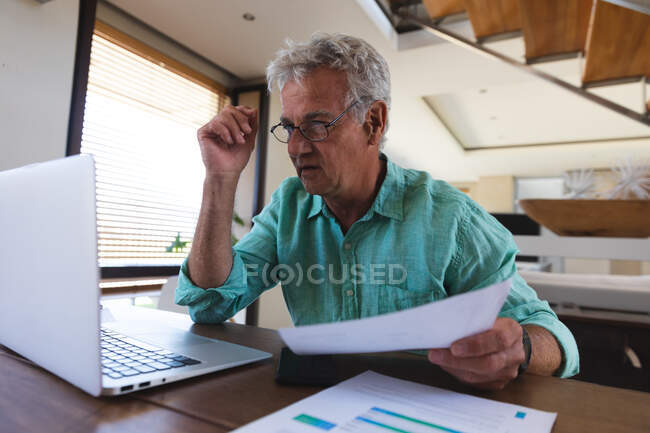Hombre caucásico mayor sentado en la mesa usando laptop pagando facturas. en casa en autoaislamiento durante coronavirus covid 19 pandemia de bloqueo - foto de stock
