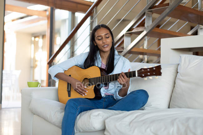 Gemischte Rassenfrau, die zu Hause auf der Couch sitzt und Gitarre spielt. Selbstisolation während der Covid 19 Coronavirus-Pandemie. — Stockfoto