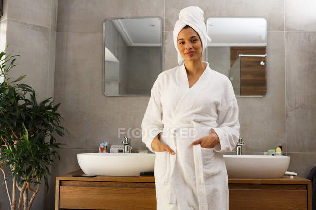Portrait de femme souriante métissée portant peignoir et serviette sur la tête debout dans la salle de bain. auto-isolement à la maison pendant la pandémie de coronavirus covid 19. — Photo de stock