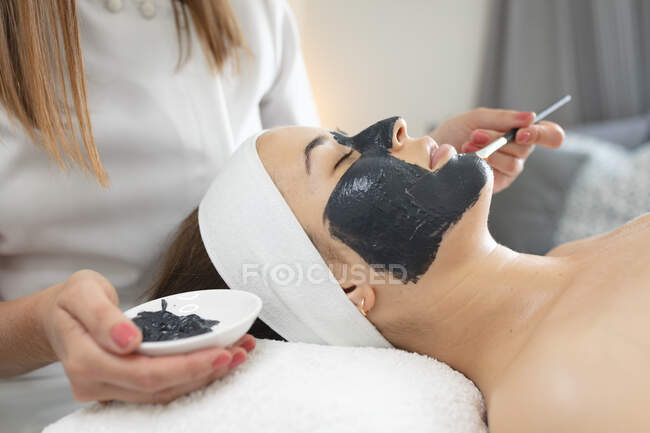 Kaukasische Frau lehnt sich zurück, während Kosmetikerin eine Gesichtsmaske aufträgt. Kundin genießt Behandlung im Schönheitssalon. — Stockfoto