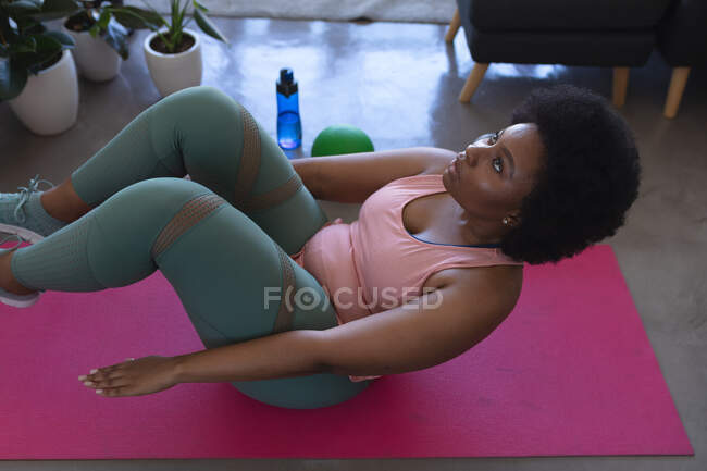 Mujer afroamericana acostada en una alfombra de ejercicio haciendo ejercicio. autoaislamiento fitness en casa durante coronavirus covid 19 pandemia. - foto de stock