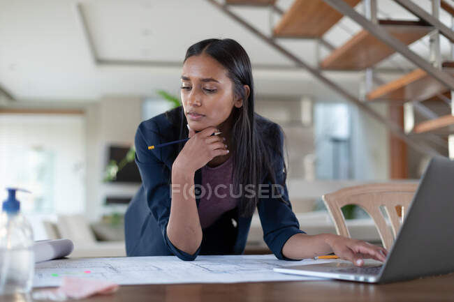 Mulher de raça mista sentada à mesa usando laptop trabalhando em casa. auto-isolamento durante a pandemia do coronavírus covid 19. — Fotografia de Stock