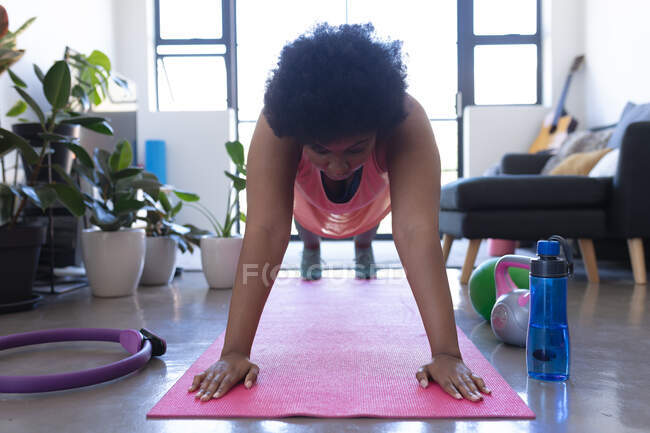 Mujer afroamericana apoyada en una colchoneta de ejercicio. autoaislamiento fitness en casa durante coronavirus covid 19 pandemia. - foto de stock