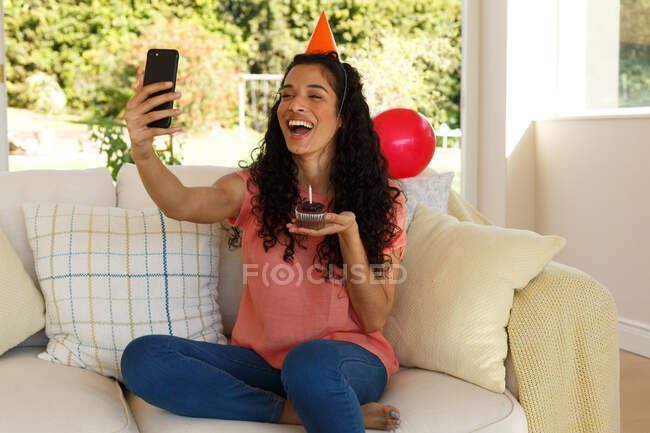 Mixed Race Frau feiert Geburtstag mit Video-Chat auf dem Smartphone. Sie trägt einen Partyhut und hält einen Muffin mit Kerze darauf. Selbstisolation zu Hause während der Covid 19 Coronavirus-Pandemie. — Stockfoto