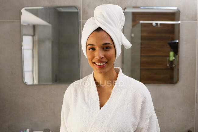 Portrait of smiling mixed race woman wearing bathrobe and towel on head in bathroom. self isolation at home during covid 19 coronavirus pandemic. — Stock Photo