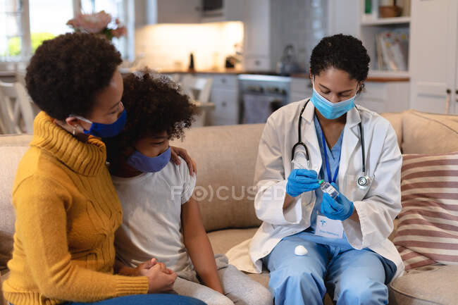 Смешанная расовая девочка и мать в масках для лица с врачом смешанной расы, готовящей вакцину против гриппа. самоизоляция дома во время пандемии коронавируса 19. — стоковое фото