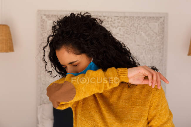 Donna di razza mista che indossa una maschera per il viso che tossisce nel braccio. auto isolamento a casa durante covid 19 coronavirus pandemia. — Foto stock