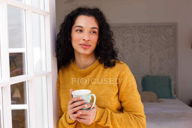 Smiling mixed race woman holding cup leaning and looking away. self isolation at home during covid 19 coronavirus pandemic. — Stock Photo