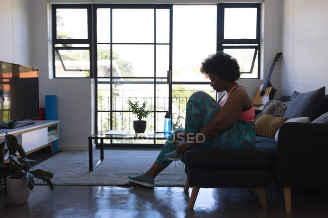 Uma afro-americana sentada no sofá a calçar sapatos desportivos. auto-isolamento em casa durante coronavírus covid 19 pandemia. — Fotografia de Stock