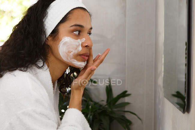 Donna razza mista guardando nello specchio applicando crema viso in bagno. auto isolamento a casa durante covid 19 coronavirus pandemia. — Foto stock