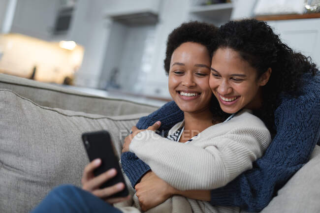 Lesbisches Paar mit gemischter Rasse sitzt mit Smartphone auf der Couch. Selbst-Isolation Qualität Familienzeit zu Hause zusammen während Coronavirus covid 19 Pandemie. — Stockfoto