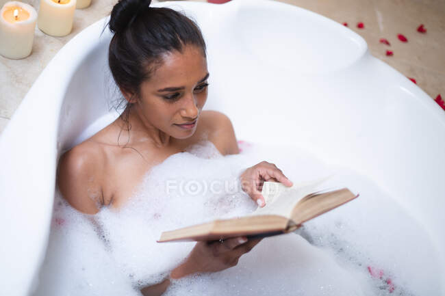 Mixed race woman lying in bath relaxing and reading book. self isolation during covid 19 coronavirus pandemic. — Stock Photo