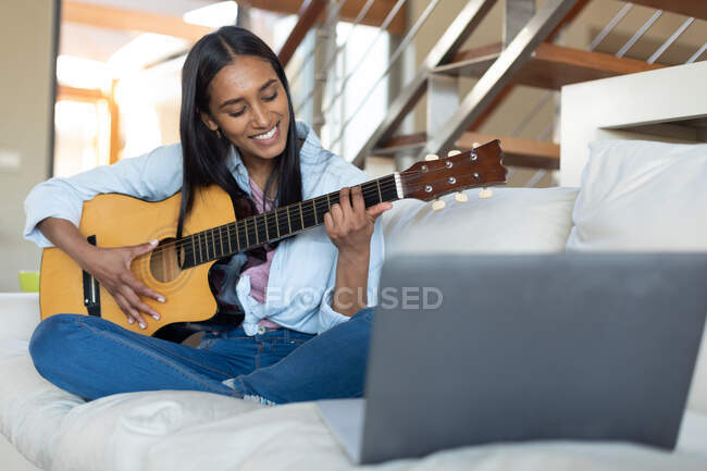 Sorrindo mulher de raça mista sentado no sofá tocando guitarra em casa. auto-isolamento durante a pandemia do coronavírus covid 19. — Fotografia de Stock