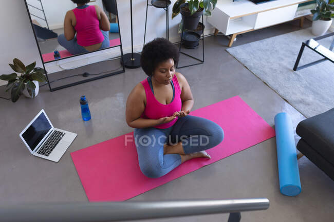 Donna afroamericana che fa meditazione yoga seduta su un tappeto indossando vestiti sportivi. computer portatile in background. auto isolamento fitness benessere tecnologia a casa durante coronavirus covid 19 pandemia. — Foto stock