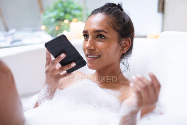 Mujer de raza mixta tumbada en el baño relajante y el uso de smartphone. autoaislamiento durante la pandemia de coronavirus covid 19. - foto de stock