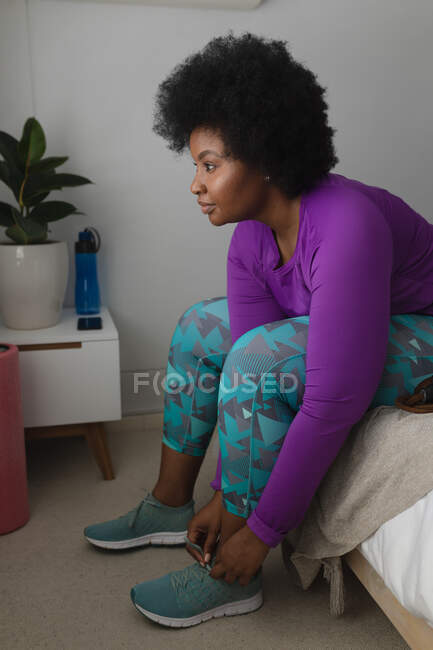 Mujer afroamericana poniéndose ropa deportiva en el dormitorio. autoaislamiento en casa durante la pandemia del coronavirus covid 19. - foto de stock