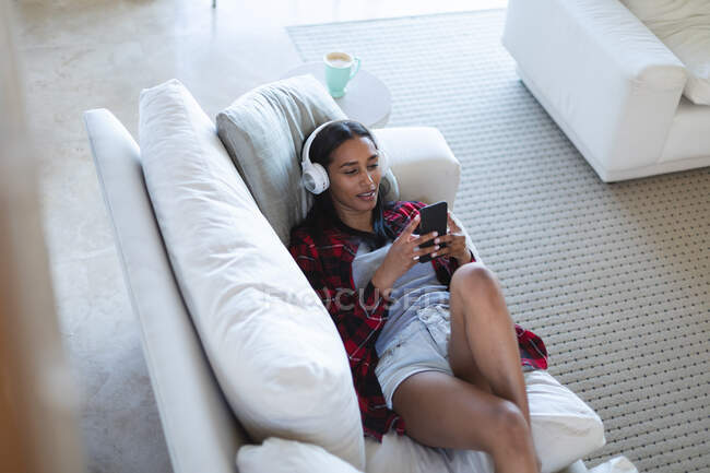 Смешанная расовая женщина лежит на диване с наушниками, используя смартфон дома. самоизоляция во время пандемии 19 коронавируса. — стоковое фото