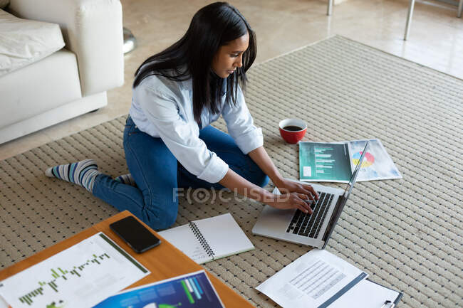 Femme de race mixte assis sur le sol en utilisant un ordinateur portable travaillant à la maison. auto-isolement pendant la pandémie de coronavirus covid 19. — Photo de stock