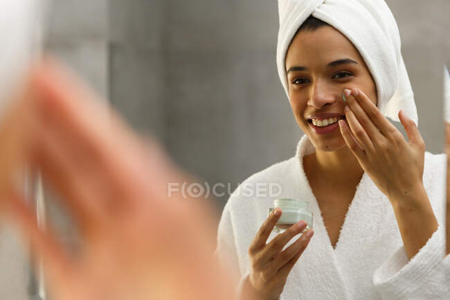 Mixed race woman looking in mirror applying face cream in bathroom. self isolation at home during covid 19 coronavirus pandemic. — Stock Photo