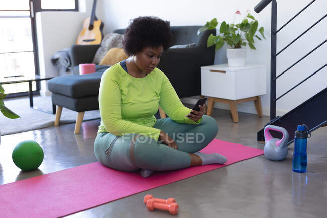 Afroamerikanerin sitzt mit Smartphone auf Gymnastikmatte Selbst-Isolation Fitness-Technologie Kommunikation zu Hause während Coronavirus covid 19 Pandemie. — Stockfoto