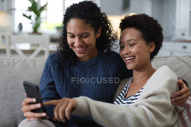 Смешанная раса лесбиянок пара, сидящая на диване с помощью смартфона. самоизоляция качество семейное время дома вместе во время коронавируса ковид 19 пандемии. — стоковое фото