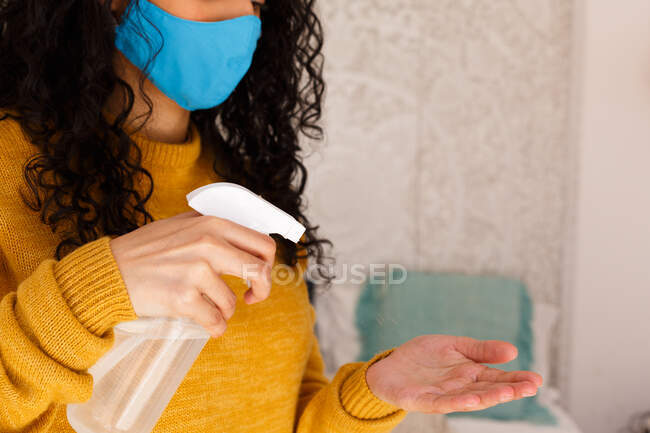 Mischlingshündin mit Mundschutz desinfiziert Hände mit Spray. Porträt einer Frau gemischter Rasse mit langen Haaren, die Gesichtsmaske trägt. Selbstisolation zu Hause während der Covid 19 Coronavirus-Pandemie. — Stockfoto