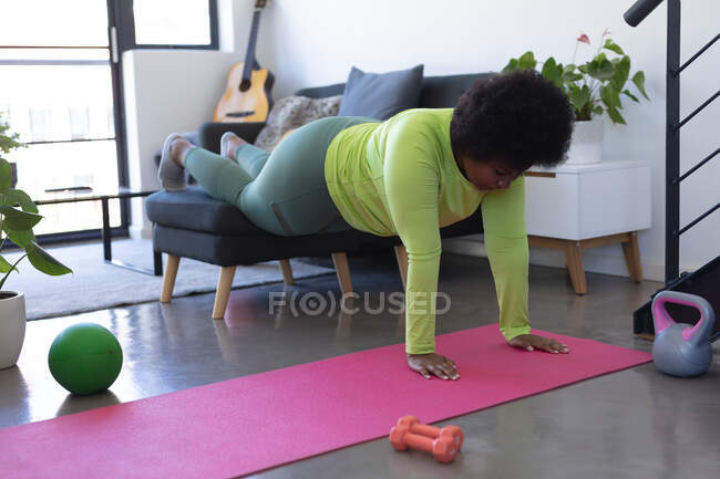 Mujer afroamericana usando silla y colchoneta de ejercicio haciendo ejercicio. autoaislamiento fitness en casa durante coronavirus covid 19 pandemia. - foto de stock