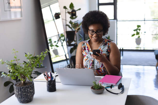 Mujer afroamericana sonriente sentada en el escritorio usando smartphone y laptop. auto aislamiento tecnología comunicación en el hogar durante coronavirus covid 19 pandemia. - foto de stock