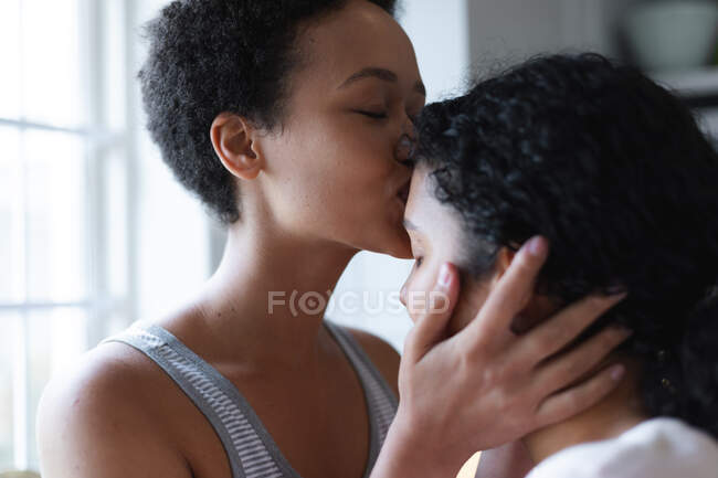 Gemischte Rasse lesbisches Paar küsst sich in der Küche. Selbstisolierung Qualität Zeit zu Hause zusammen während Coronavirus covid 19 Pandemie. — Stockfoto