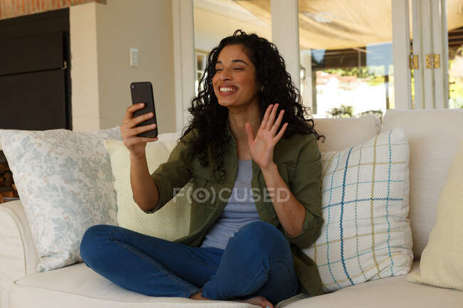Змішана расова жінка, яка махає відео чатом на смартфоні. сидячи на дивані у вітальні. самоізоляція в домашніх умовах під час пандемії коронавірусу 19 . — стокове фото