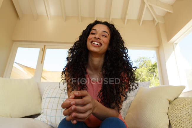 Gemischte Rasse Frau sitzt auf Couch im Wohnzimmer. in die Kamera schauend und lächelnd. Selbstisolation während der Covid 19 Coronavirus-Pandemie — Stockfoto