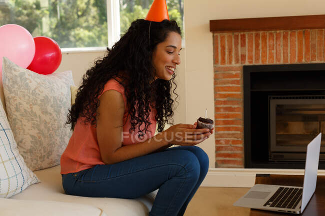 Mujer de raza mixta celebrando cumpleaños teniendo chat de vídeo en el ordenador portátil. llevando sombrero de fiesta y sosteniendo panecillo con vela. autoaislamiento en el hogar durante la pandemia de coronavirus covid 19. - foto de stock