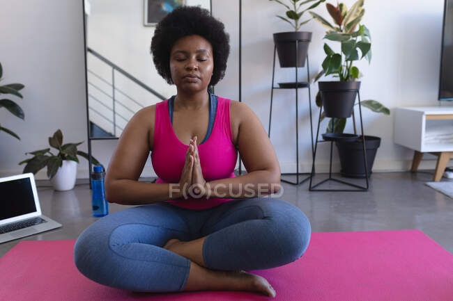 Mujer afroamericana meditando sentada en una alfombra con ropa deportiva. portátil en el fondo. autoaislamiento fitness bienestar tecnología en el hogar durante coronavirus covid 19 pandemia. - foto de stock