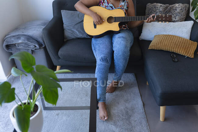 Mujer afroamericana sentada en un sofá tocando la guitarra acústica. autoaislamiento hobby tiempo música en casa durante coronavirus covid 19 pandemia. - foto de stock