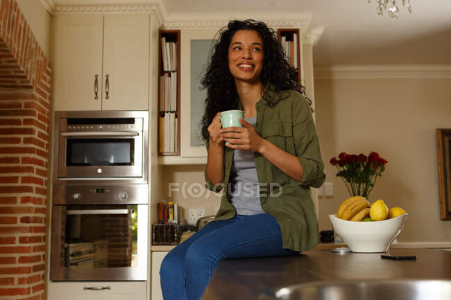 Mixed race woman holding cup of coffee sitting in kitchen. self isolation at home during covid 19 coronavirus pandemic. — Stock Photo