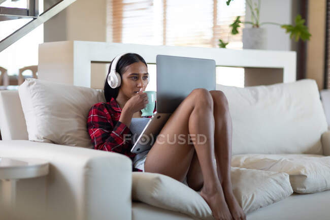 Donna razza mista con cuffie sul computer portatile, avendo caffè sul divano a casa. autoisolamento durante la pandemia di covid 19 coronavirus. — Foto stock