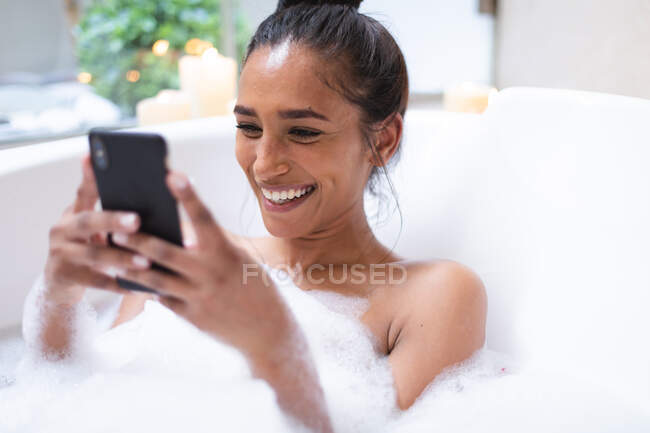 Mulher de raça mista deitada no banho relaxante e usando smartphone. auto-isolamento durante a pandemia do coronavírus covid 19. — Fotografia de Stock