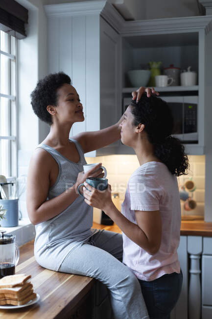 Smiling mixed race lesbian couple having coffee and talking in kitchen. self isolation quality time at home together during coronavirus covid 19 pandemic. — Stock Photo