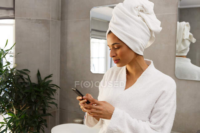 Жінка змішаної раси носить халат і рушник на голові, використовуючи смартфон у ванній кімнаті. самоізоляція в домашніх умовах під час пандемії коронавірусу 19 . — стокове фото
