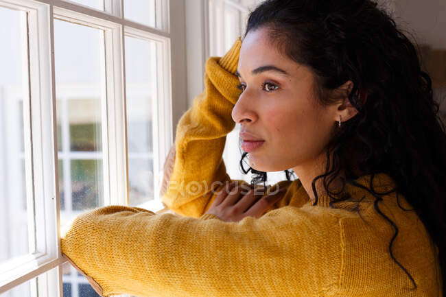 Mulher de raça mista de pé e olhando pela janela. auto-isolamento em casa durante a pandemia do coronavírus covid 19. — Fotografia de Stock