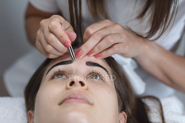 Kaukasische Frau lehnt sich zurück, während Kosmetikerin ihre Augenbrauen zupft. Kundin genießt Behandlung im Schönheitssalon. — Stockfoto