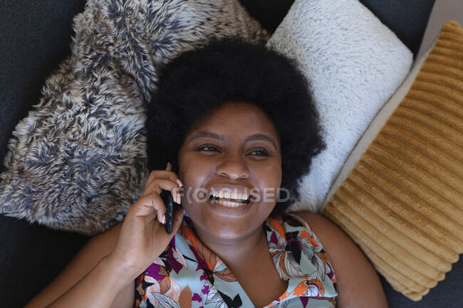 Sorrindo afro-americana deitada no sofá usando um smartphone. comunicação de tecnologia de auto-isolamento em casa durante a pandemia do coronavírus covid 19. — Fotografia de Stock