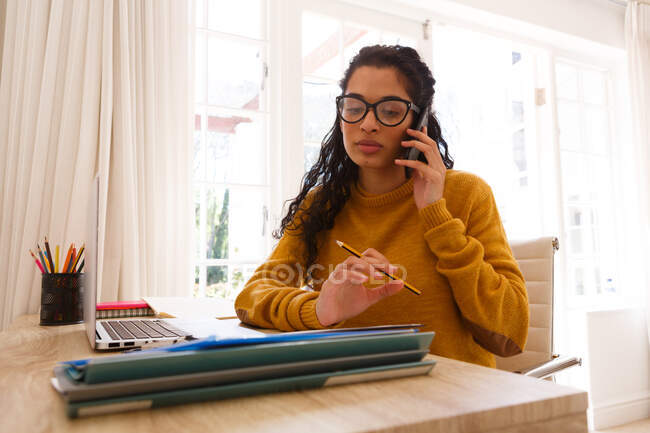 Смешанная расовая женщина разговаривает на смартфоне, сидя за столом с ноутбуком и бумажной работой. самоизоляция в домашних условиях во время пандемии 19 коронавируса. — стоковое фото
