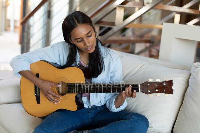Mixed race woman sitting on couch playing guitar at home. self isolation during covid 19 coronavirus pandemic. — Stock Photo