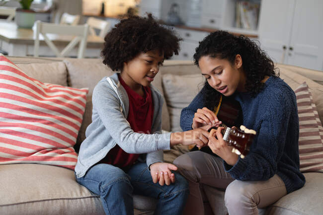 Mixed race woman and daughter sitting on couch. playing a guitar. self isolation quality family time at home together during coronavirus covid 19 pandemic. — Stock Photo