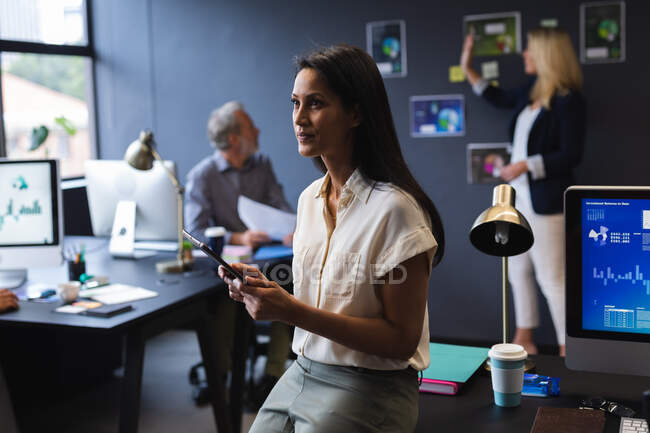 Деловая женщина смешанной расы сидит на столе, используя цифровой планшет в креативном офисе. современные офисные технологии для совместной работы. — стоковое фото