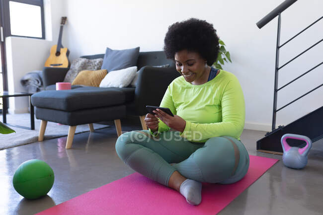 Mujer afroamericana sentada en una colchoneta de ejercicio usando un teléfono inteligente. auto aislamiento fitness tecnología comunicación en el hogar durante coronavirus covid 19 pandemia. - foto de stock