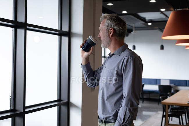 Homem de negócios caucasiano à janela a beber café no escritório moderno. negócio moderno escritório tecnologia do local de trabalho. — Fotografia de Stock