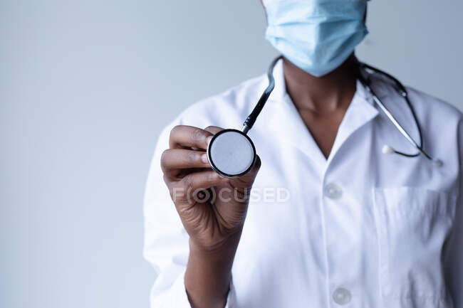 Médico femenino de raza mixta con máscara facial de pie y usando un estetoscopio. higiene profesional médica del trabajador sanitario durante la pandemia del coronavirus covid 19. - foto de stock