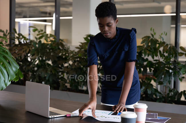 Змішана гоночна бізнес-леді стоїть за допомогою ноутбука, що проходить через паперові роботи в сучасному офісі. бізнес сучасні технології офісного робочого місця . — стокове фото